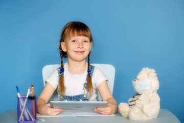 A girl 5-6 years old sitting at a table performs homework remote tasks on the tablet. Isolate on a blue background.