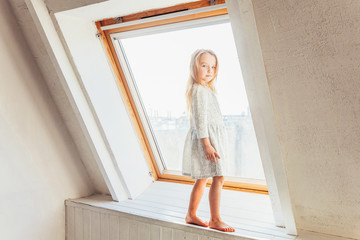 Stay Home Stay Safe. Little cute sweet smiling girl in white dress standing on window sill in bright light living room at home indoors. Childhood schoolchildren youth relax concept.