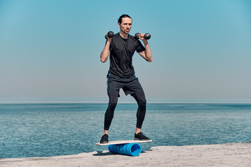 Closeup Of Healthy Handsome Active Man With Fit Muscular Body keeping balance with dumbbells on the wooden board against the background of sea on summer day