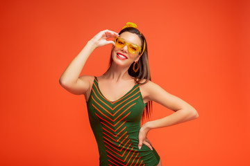 Happy woman with hand on hip touching sunglasses, smiling and looking at camera isolated on orange