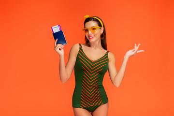 Front view of woman smiling, looking at camera and showing passport with air ticket isolated on orange