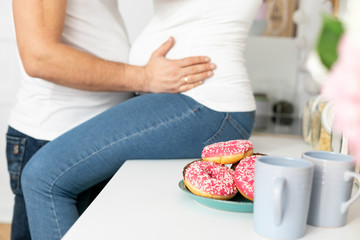 Obraz na płótnie Canvas Man holding belly of his pregnant wife, sitting on the table in the kitchen with donuts in front