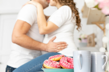 Obraz na płótnie Canvas Man holding belly of his pregnant wife and kisses, sitting on the table in the kitchen with donuts in front