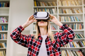 Pretty Caucasian student girl, wearing casual chekered shirt, using VR glasses for work and studying in university library. VR technology concept and education