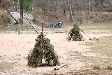 Two tepees built with twigs and branches of conifers standing on sandy ground in the forest with bycicles in the background Seen in Germany in April.