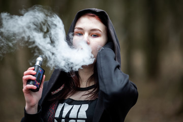 Vape teenager. Young pretty caucasian brunette girl with problem skin in the hood smoking an...