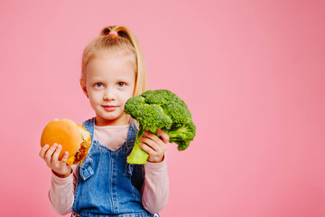 Young smiling blonde girls is holding a hamburger and broccoli on pink background. The choice...