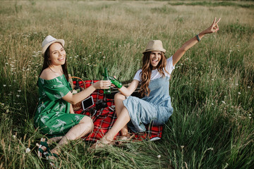 Two attractive young women sitting down on the picnic blanket and have fun. Beer and sandwiches.