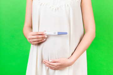 Obraz na płótnie Canvas Close up of pregnant woman in white dress holding positive pregnancy test at green background. Motherhood concept. Copy space