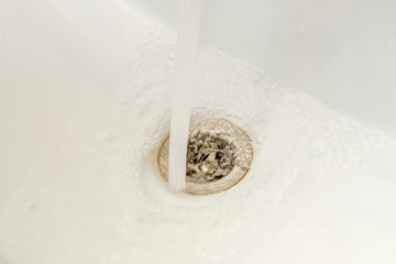 Water from the tap pours into a white ceramic sink. Drain water in the sink close up