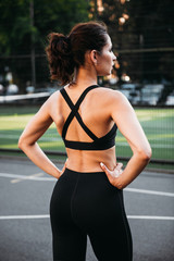 Fototapeta premium Beautiful sexy fitness young woman wearing black leggings and sport bra standing at the court outside. Energetic fit caucasian dark hair girl posing for the camera.