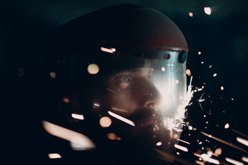 Adult bearded man in transparent protective mask and grinder saw with flying metal particles sparks in darkness