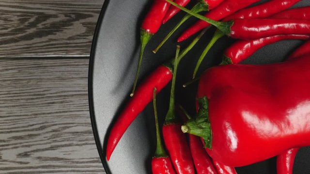 Top View Of Fresh Raw Red Chili Peppers And Ramiro Pepper Rotating On A Black Plate On A Gray Wooden Background On The Right Side.