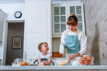 A smiling girl looks at her mother, who is beating the dough with a mixer