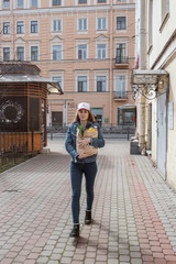 Smiling girl in cap and denim clothes walking with kraft brown paper bag with fresh vegetables and food in hands. Delivery service from shop or restaurant concept. Young woman grocery courier. Covid19