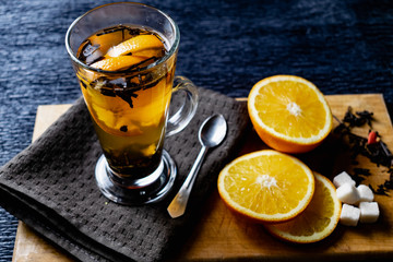 Citrus drink, tea with petals in a transparent glass mug with slices of sweet orange and refined sugar. Slicing fruit on a wooden Board with a brown napkin in a cozy dark background in a low key