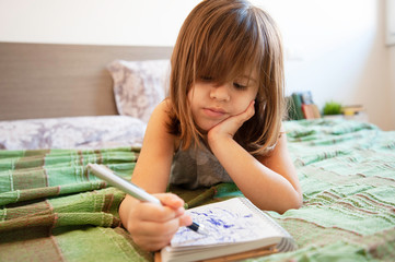 Little cute preschool girl lying on the bed in bedroom at home, she's bored and scribbling with blue pen on the paper notebook. Childhood concept. Leisure activity indoors. Getting bored at home.