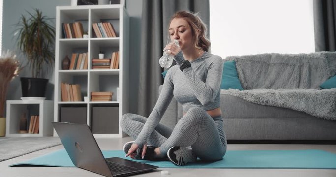Young sporty woman in activewear drinking water and looking at computer screen while resting after workout. Charming girl with blond hair sitting on yoga mat with crossed legs.