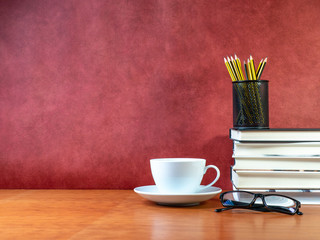 cup, books, glasses and pencil pot on wooden table and red background