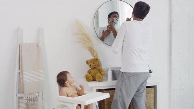 Medium shot of Indian father looking in mirror in bathroom and shaving while adorable toddler girl sitting in highchair and chewing toothbrush