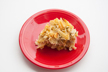 Slice of tuna noodle casserole cut and ready to eat on a red plate with a light background.  This piece of casserole has peas in it and potato chips on top.