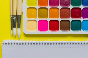 Creative layout with aquarelle palette, paint brushes and sketchbook on yellow background. Top view. Artists concept, copy space for text.