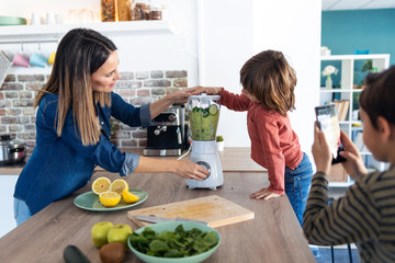 Boy helping his mother to prepare a detox juice with blender while his brother take photographs...