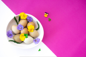 Easter eggs with yellow and lilac flowers in a white bowl on a white and pink background.