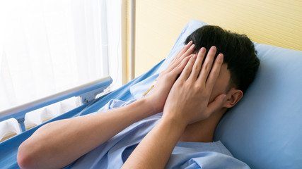 Asian patient young male anxious, stress and sensitive feeling from quarantine so raise hand palm to cover face on his bed in sickroom after admitted at hospital for long days