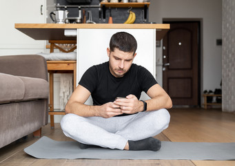 man in sportswear sitting on a yoga mat in the interior of the apartment is studying lessons watching online broadcast on a smartphone. Home fitness