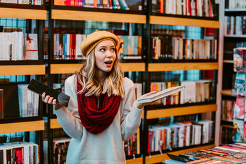 Portrait of attractive blonde woman posing next to shelves with books at book store. Tablet or book decision. 