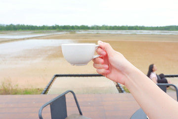 Fototapeta na wymiar Hand women hodling white coffee cup with salt farm background. She hand keep cup for drinking coffee for relaxing on holiday time.CGirl is holding white cup in hands. Mug for woman, gift.Close up.
