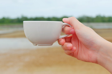 Hand women hodling white coffee cup with salt farm background. She hand keep cup for drinking coffee for relaxing on holiday time.CGirl is holding white cup in hands. Mug for woman, gift.Close up.