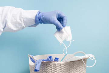 a hand in a blue disposable medical glove throws a used protective face mask into a trash can, blue...