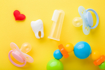 Red heart, soothers, white tooth and transparent silicone fingertip toothbrush and colorful rattle on bright yellow table background. Things for babies teeth cleaning through fun. Close up. Top view.