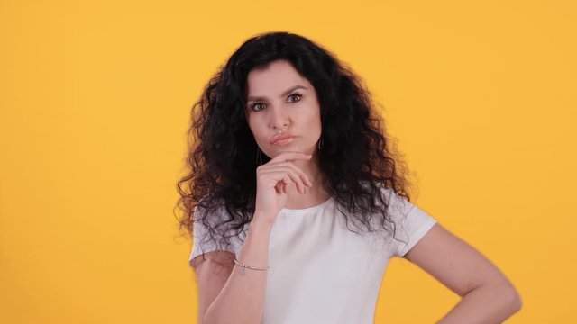 Portrait of beautiful young woman with black curly hair is thoughtful doesnt know undecided isolated on yellow background shot in 4k