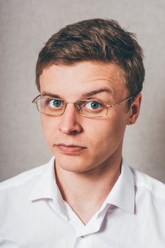 Man Serious In Glasses Raised One Eyebrow Looking At The Camera. On A Gray Background.