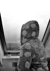 black and white photo of woman in hooded dressing gown looking out of rainy window 
