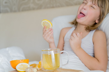 Sick girl in bed tastes lemon slice.Cup of antipyretic drugs for colds,flu.Tea with citrus vitamin C,ginger root,orange.Wooden tray.Home self-treatment.Medical quarantine covid-19 coronavirus therapy