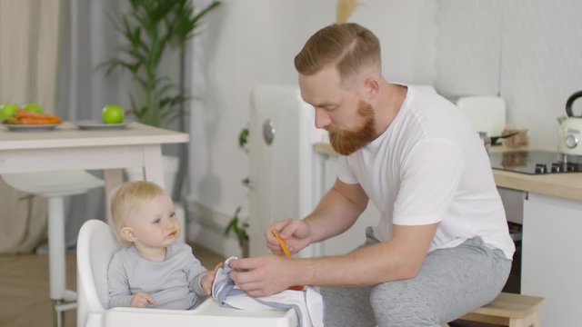 Medium shot of bearded father feeding adorable baby girl sitting in highchair in kitchen