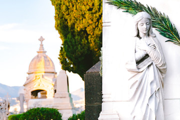 sculpture of a saint maria woman with crossed hands in an old cemetery. stoned statue praying at cemetery. Graveyard old weathered stone sad mother statue on funeral. church, mountains on background