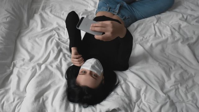 Young woman take selfie or make video call in protective medical face mask at home or hospital lying on white bed. Self-isolation and quarantine, epidemic or pandemic of covid-19 coronavirus