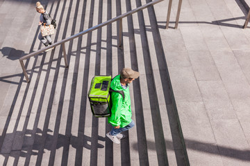 From above side view shot of young male food courier in green uniform witn insulated bag walking upstairs leaving subway while delivering order