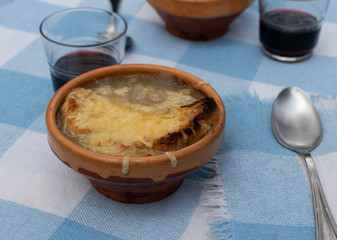 Plates of traditional French gratin onion soup on a rustic table with blue squared tablecloth, spoons and ceramic plates with glasses of wine.