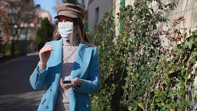 Woman wears reusable mask outdoors during coronavirus covid-19 pandemic. Girl using smartphone on empty street. Stay safe. Spring fashion