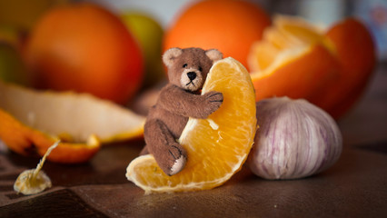 A small toy bear hugs a slice of orange against a background of fruit and garlic