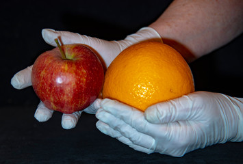 Women's hands in rubber gloves hold an Apple and an orange on a black background.