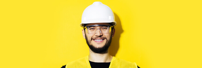 Studio panoramic portrait of young smiling construction engineer worker man, wearing safety helmet and goggles on background of yellow color with copy space.