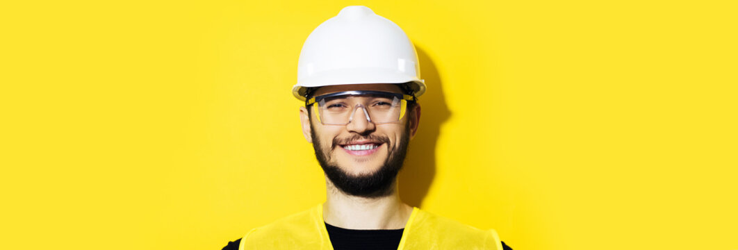 Studio Panoramic Portrait Of Young Smiling Construction Engineer Worker Man, Wearing Safety Helmet And Goggles On Background Of Yellow Color With Copy Space.