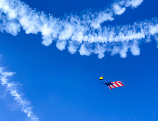 Stunt Skydiver with Parachute and an American Flag against Blue Sky with Smoke Airplane Contrails